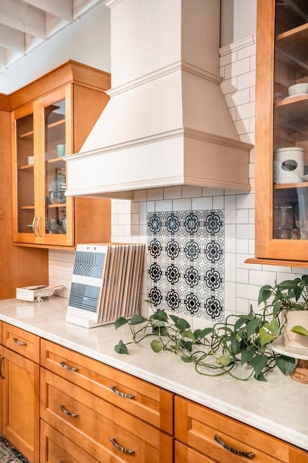 A modern kitchen with light wood cabinets, white countertops, a white range hood, black and white patterned tile backsplash, and a green trailing plant on the counter—see this style at our Spencerport Showroom.