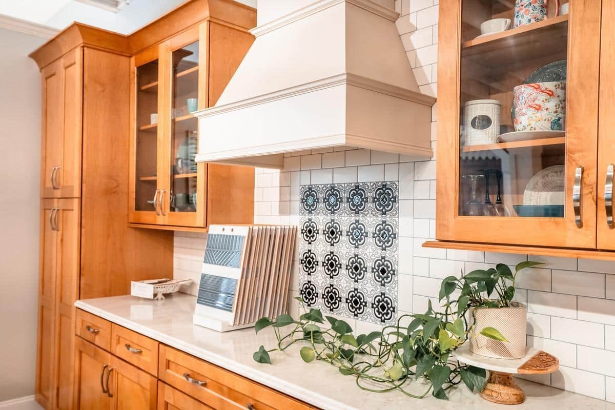 A bright kitchen in the Spencerport Showroom features wooden cabinets, a white range hood, patterned black and white tile backsplash, white subway tiles, dishes behind glass doors, a trailing potted plant, and countertop material samples on the counter.