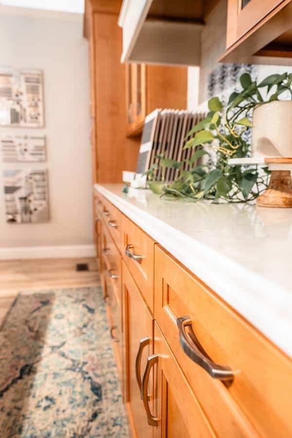 A close-up view of a kitchen with light wood cabinets, silver handles, and a white countertop from the Spencerport Showroom. A green plant sits nearby, while a patterned rug and framed wall art add charm to the wooden floor in the background.