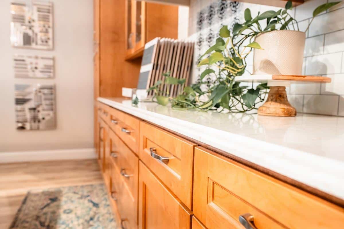 Close-up of a kitchen counter with light wood cabinets in the Spencerport Showroom, a white countertop, a green potted plant, cutting boards, and kitchen tiles in the background. A rug and wall art are visible in the background.