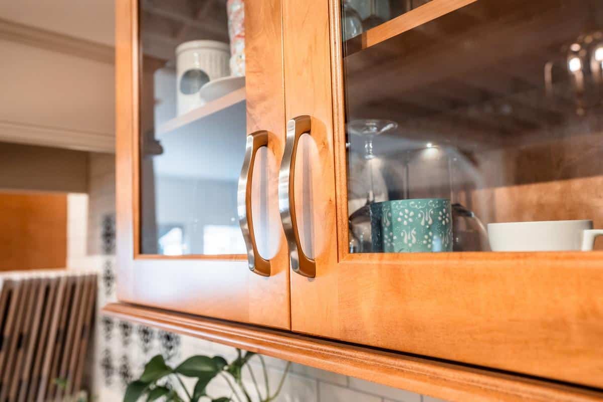 Close-up of wooden kitchen cabinet doors with glass panels in a Spencerport showroom, showing reflections and various cups and dishes inside. The handles are metallic and curved, with part of a plant and tiled wall visible in the background.