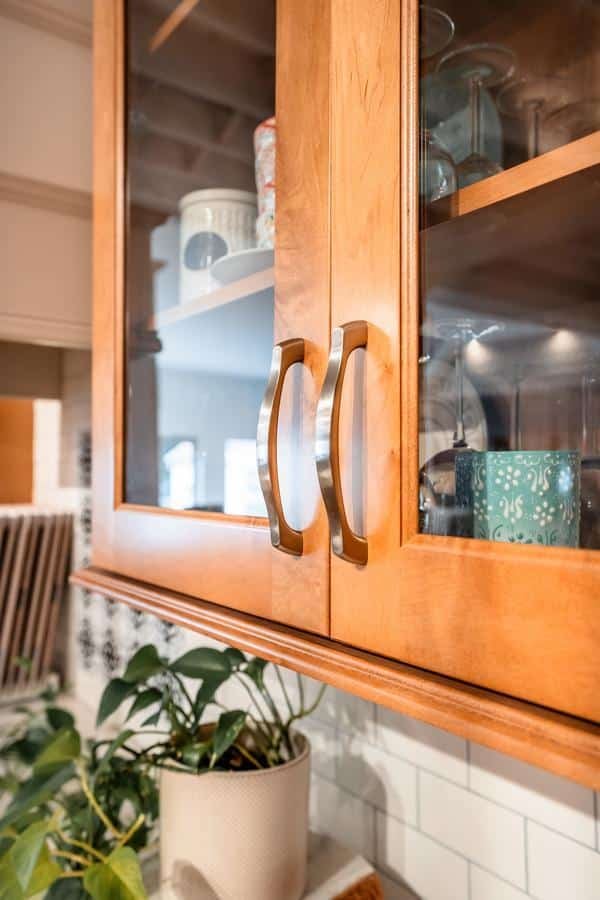 A close-up of wooden kitchen cabinet doors with glass panels, metal handles, and shelves displaying dishes and glassware in the Spencerport Showroom; a potted plant sits on the countertop below.