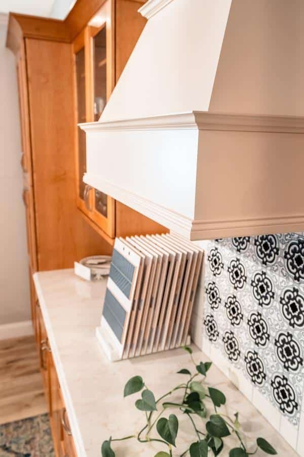 White kitchen range hood above a marble countertop in the Spencerport Showroom, with wooden cabinets, a black-and-white patterned backsplash, a file organizer, and a leafy green plant in the foreground.