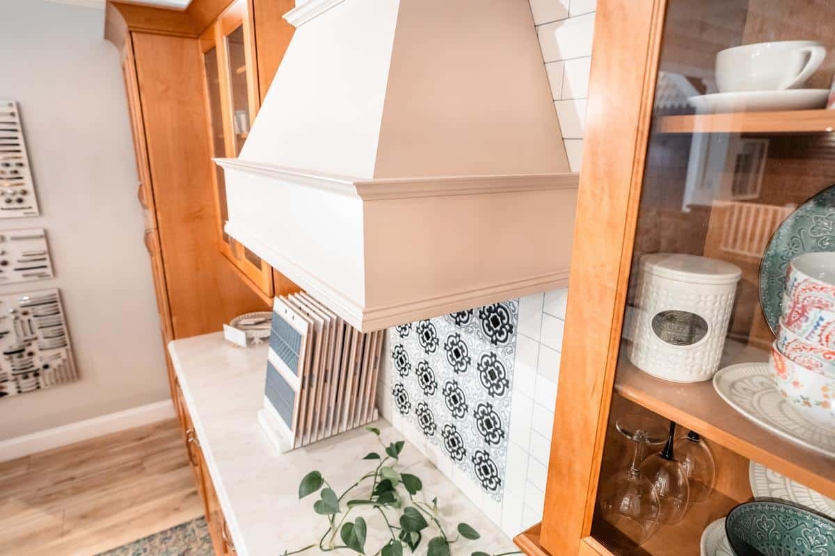 A bright kitchen features a white range hood, patterned tile backsplash, marble countertop with a plant, cookbooks, and a glass-front cabinet displaying dishes and mugs—all showcased in the inviting Spencerport Showroom. Warm wood cabinetry adds contrast.