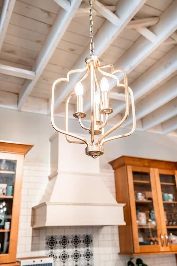 A gold chandelier with four candle-style bulbs hangs from the ceiling in a kitchen with exposed white beams, wooden cabinets, and a white subway tile backsplash—find similar styles at the Spencerport Showroom.