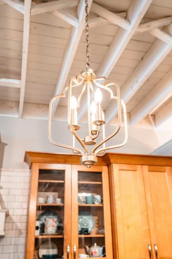A gold chandelier with three candle-style light bulbs hangs from a chain in a kitchen with wooden cabinets, white subway tile, and an exposed wooden beam ceiling—find inspiration at the Spencerport Showroom.