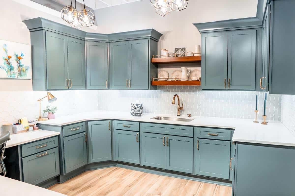 Modern kitchen with teal cabinets, white countertops, stainless steel sink, wooden floating shelf with dishes, gold faucet, geometric pendant lights, and light wood flooring—featured in our Spencerport Showroom.