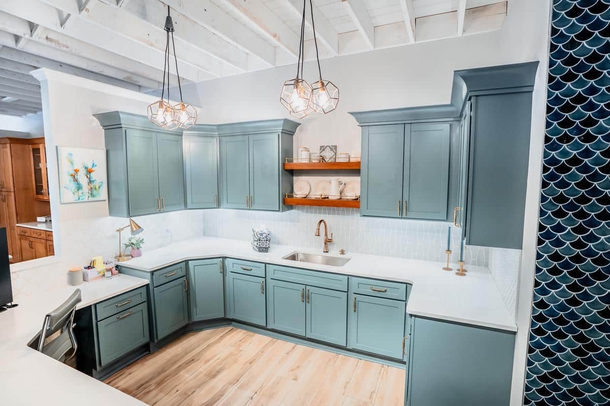 Modern kitchen with teal cabinets, white countertops, wood shelves with dishes, and geometric pendant lights. The Spencerport Showroom features a backsplash of white tiles and blue fish scale accents beneath exposed white beams.