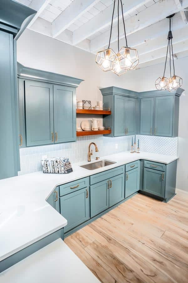 Modern kitchen with blue cabinets, white countertops, open wooden shelves, geometric pendant lights, and light wood flooring. Visit our Spencerport Showroom to see the striking white geometric tile backsplash in person.