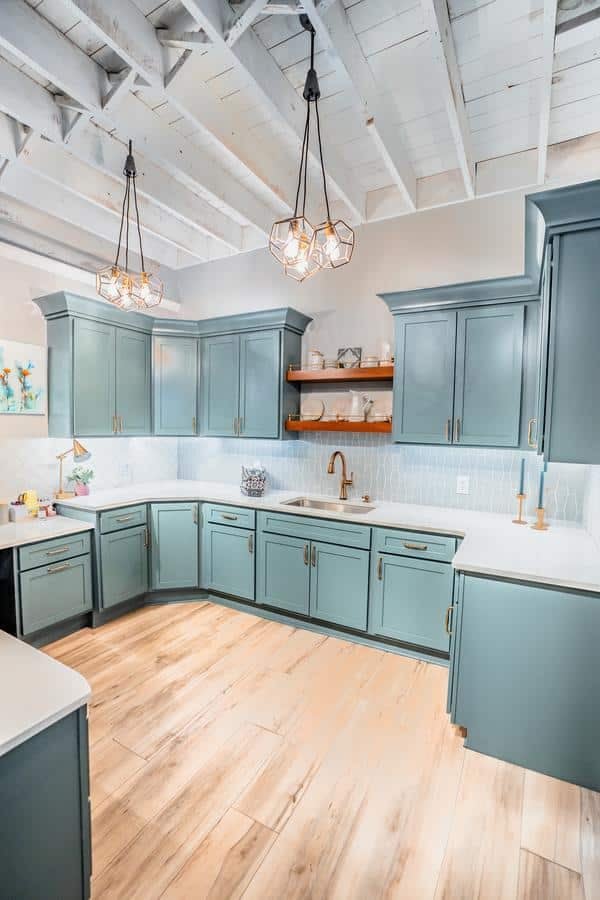 Modern kitchen with teal cabinets, white countertops, light wood floors, and open shelves displaying dishes—just like you’d find in a Spencerport showroom, complete with pendant lights and a white exposed beam ceiling.