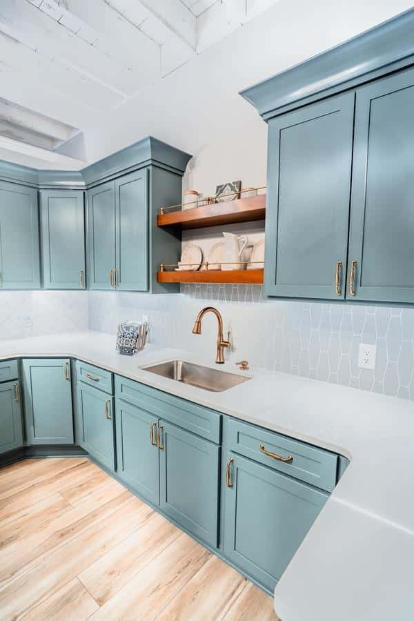 A modern kitchen with light blue cabinets, gold hardware, white countertops, a stainless steel sink, wooden shelves displaying dishes, white tile backsplash, and light wood flooring awaits you at the Spencerport Showroom.