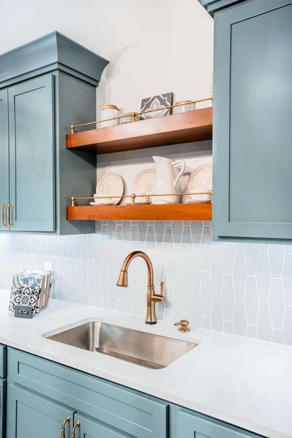 Modern kitchen with teal cabinets, gold hardware, a stainless steel sink, and a gold faucet. White countertops and a patterned backsplash add style, while wooden open shelves display dishes—perfect inspiration for your Spencerport showroom.