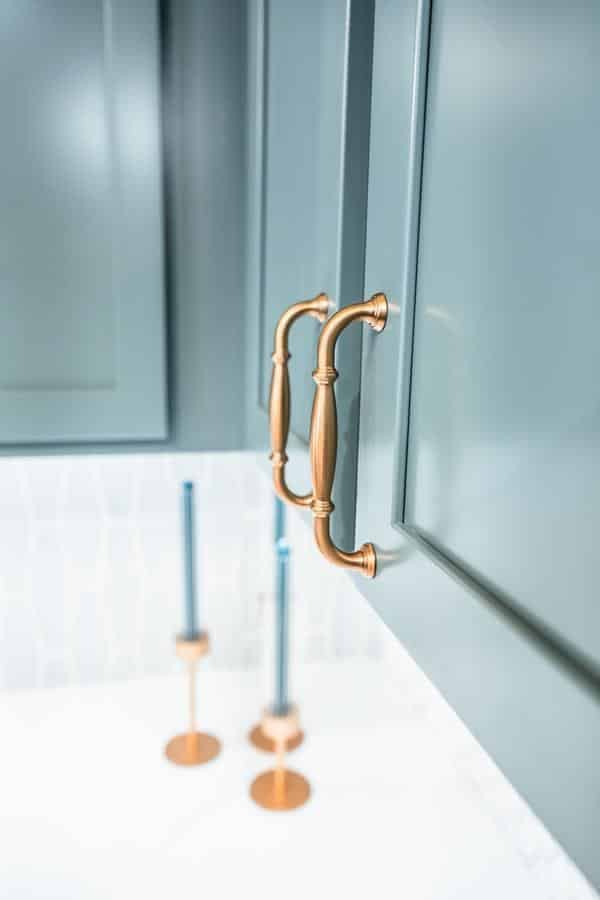 Close-up of two gold cabinet handles on teal-colored doors in the Spencerport Showroom, with blurred blue and gold candlesticks in the background on a white countertop.