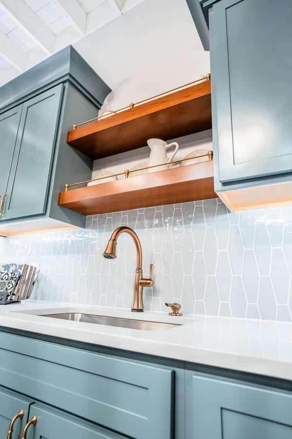 A modern kitchen with teal cabinets, white countertops, a gold faucet, and wooden open shelves displaying dishes, featuring a white geometric tile backsplash—style inspiration from the Spencerport Showroom.