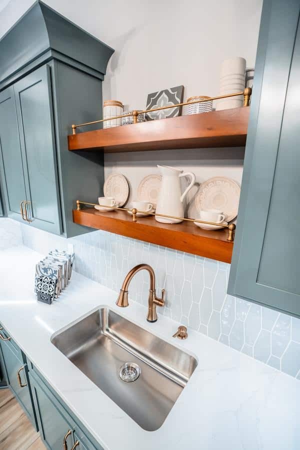 A modern kitchen sink with a gold faucet sits below wooden shelves holding white dishes, cups, and a pitcher. Green cabinets and a white countertop complement the hexagonal tile backsplash in this stylish Spencerport Showroom display.