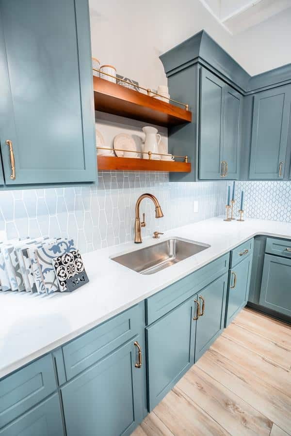 A modern kitchen showroom in Spencerport features teal cabinets with gold handles, a stainless steel sink, white countertop, geometric tile backsplash, open shelves with dishes, and light wood flooring.