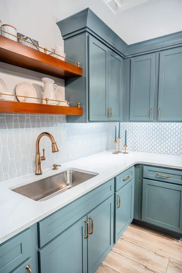 A modern kitchen from the Spencerport Showroom features teal cabinets, brass hardware, a stainless steel sink, white countertop, open wooden shelves with dishes, a geometric white tile backsplash, and light wood flooring.