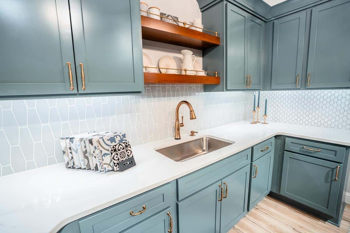 Modern kitchen with teal cabinets, brass hardware, a white countertop, and a gold faucet. Patterned cutting boards and open shelves display dishes beneath a geometric backsplash—find inspiration like this at the Spencerport Showroom.