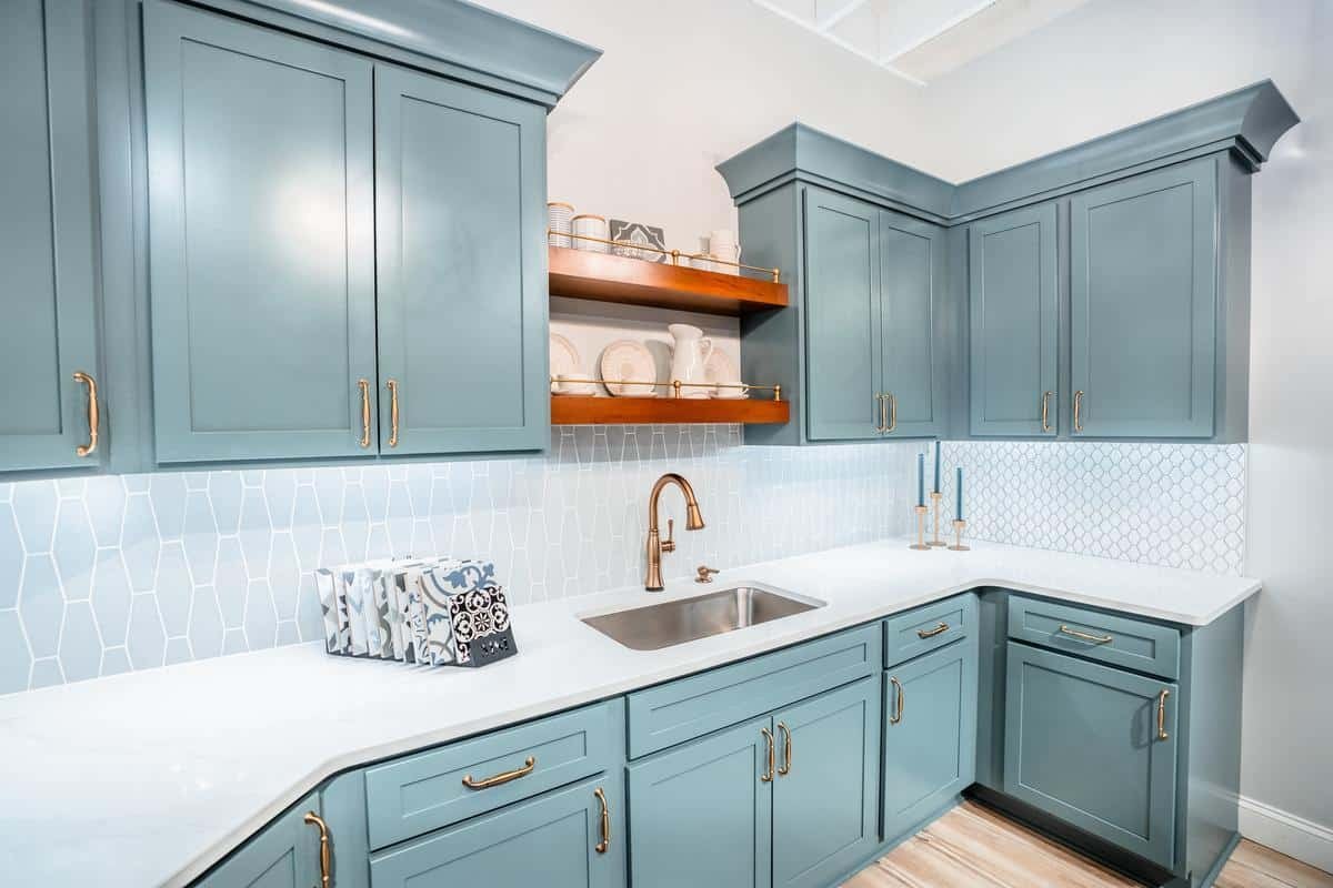 Modern kitchen with light blue cabinets, gold hardware, a white countertop, geometric tile backsplash, and stainless steel sink with gold faucet—visit our Spencerport Showroom to see wooden open shelves and light wood flooring in person.