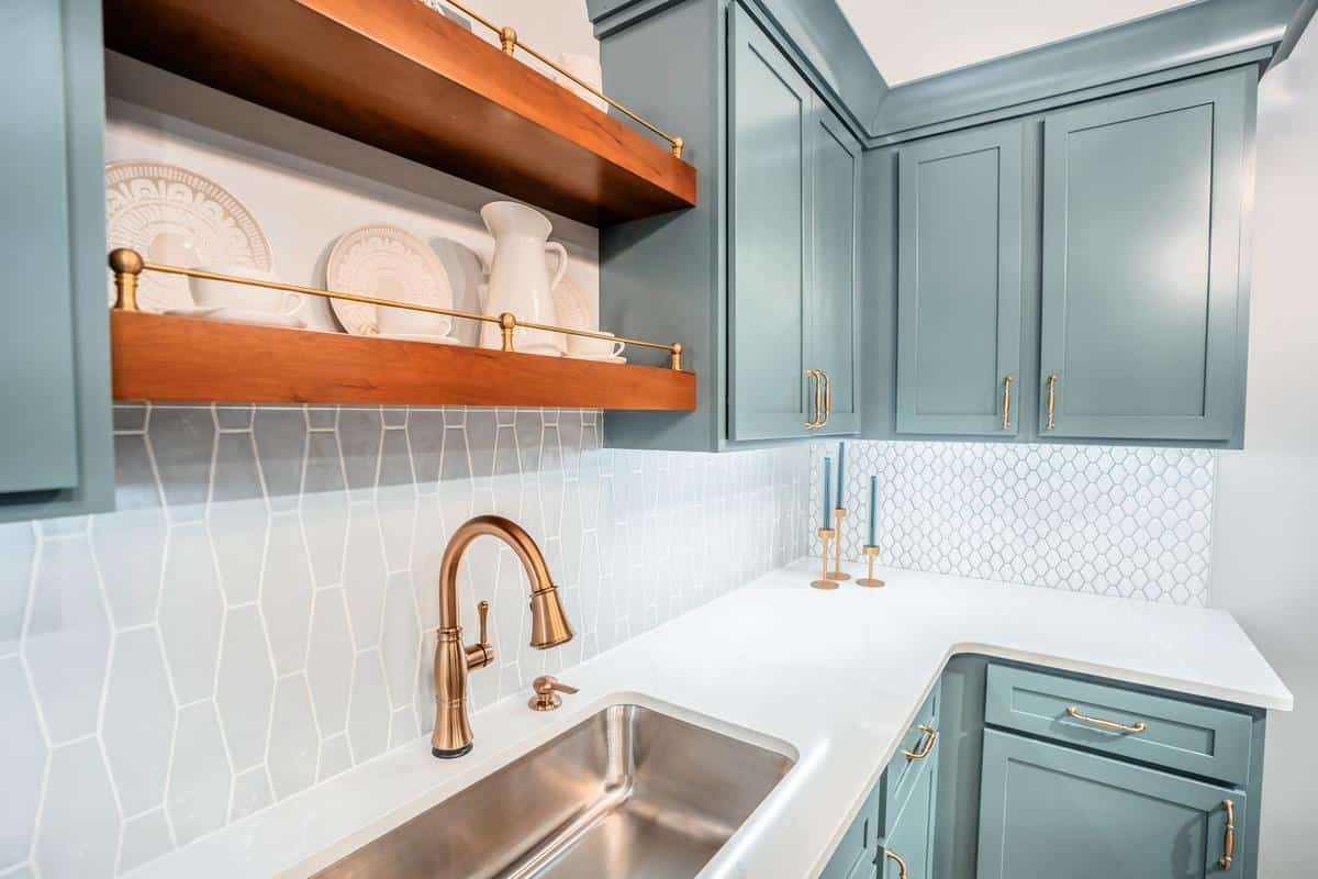 Modern kitchen with teal cabinets, gold hardware, white quartz countertop, stainless steel sink, and bronze faucet. Open wooden shelves display white dishes. Geometric tile backsplash. Visit our Spencerport Showroom to see this design in person.