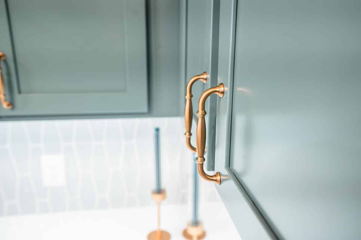 Close-up of light green cabinet doors with gold handles in a modern kitchen at the Spencerport Showroom, featuring a geometric-patterned white and gray backsplash and blurred candlesticks in the background.