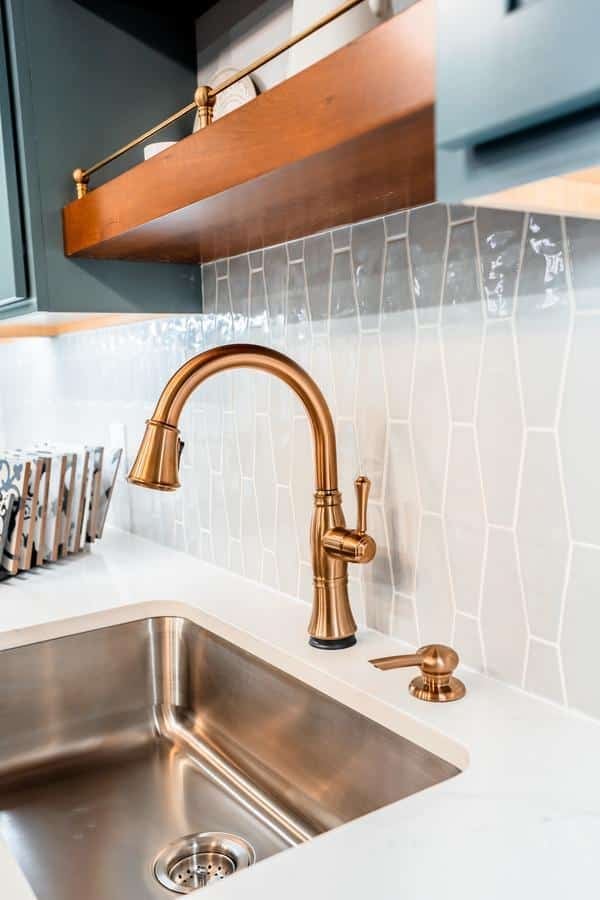 A modern kitchen sink with a brushed gold faucet and soap dispenser, set in a white countertop with a geometric white tile backsplash and wooden shelving above—discover this look at our Spencerport Showroom.