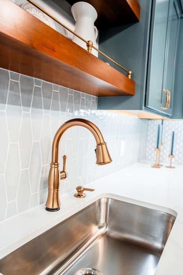 A modern kitchen sink with a sleek brushed gold faucet and soap dispenser set in a white countertop. The backsplash features geometric white tiles, while wooden shelves hold decor items above—perfect inspiration for any Spencerport showroom.