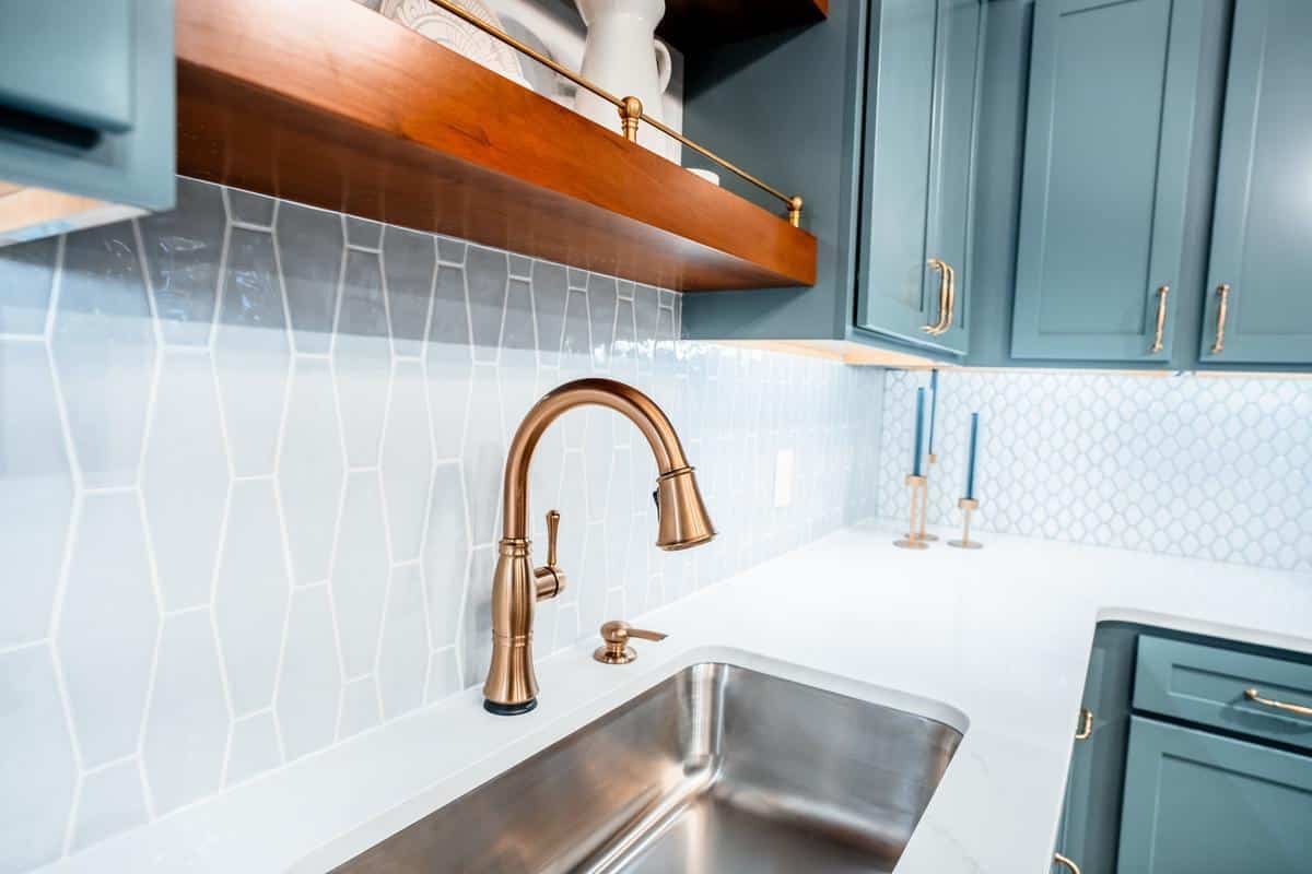 Close-up of a modern kitchen sink with a brushed gold faucet, blue cabinets, and geometric tile backsplash—styled to perfection like our Spencerport Showroom displays. Wooden open shelving and blue candles complete the sophisticated look.