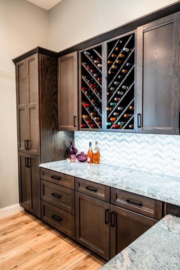 Modern kitchen with dark wood cabinets, marble countertop, chevron-patterned white backsplash, and a diagonal wine rack filled with wine bottles—just like what youll find at the Spencerport Showroom. Three bottles and two glass bowls sit on the counter.