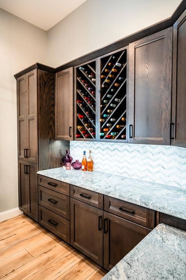 Modern kitchen showroom in Spencerport featuring dark wooden cabinets, a marble countertop, a herringbone-patterned white backsplash, and a built-in wine rack filled with bottles. Three bottles and two glass decanters sit on the counter.