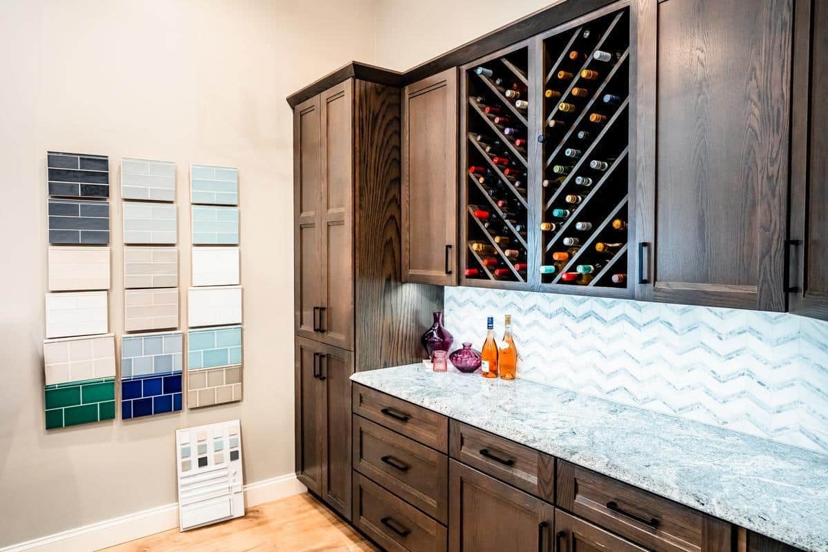 A modern kitchen display at the Spencerport Showroom features dark wood cabinets, a marble countertop, a blue chevron backsplash, a wine rack with bottles, and wall-mounted sample tiles in various colors and finishes.