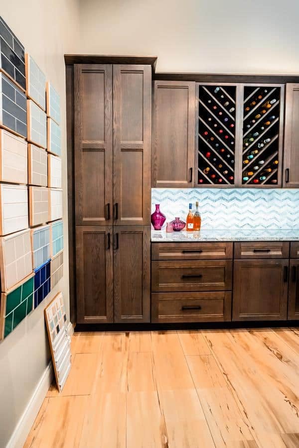 A modern showroom kitchen in Spencerport features dark wood cabinets, a wine rack filled with bottles, patterned backsplash, and sample tile boards on the wall and floor. Glass decanters and liquor bottles adorn the countertop.