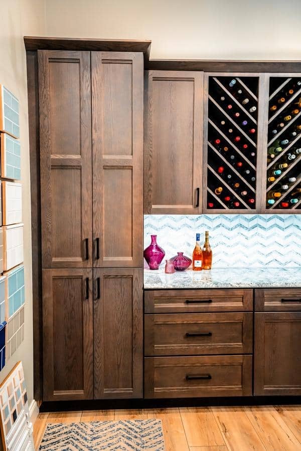 Explore our Spencerport Showroom featuring kitchen cabinets with a dark wood finish, a countertop adorned with decorative glass bottles and liquor, a zigzag tile backsplash, and a wine rack. Tile samples are beautifully displayed on the left wall.
