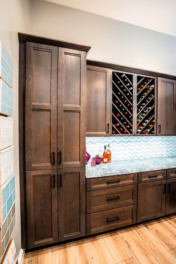 A modern kitchen in Spencerport features dark wood cabinets, a marble countertop, a chevron-patterned backsplash, and a built-in wine rack filled with bottles. Three colorful liquor bottles sit on the counter, giving it a true showroom feel. Light wood flooring completes the space.