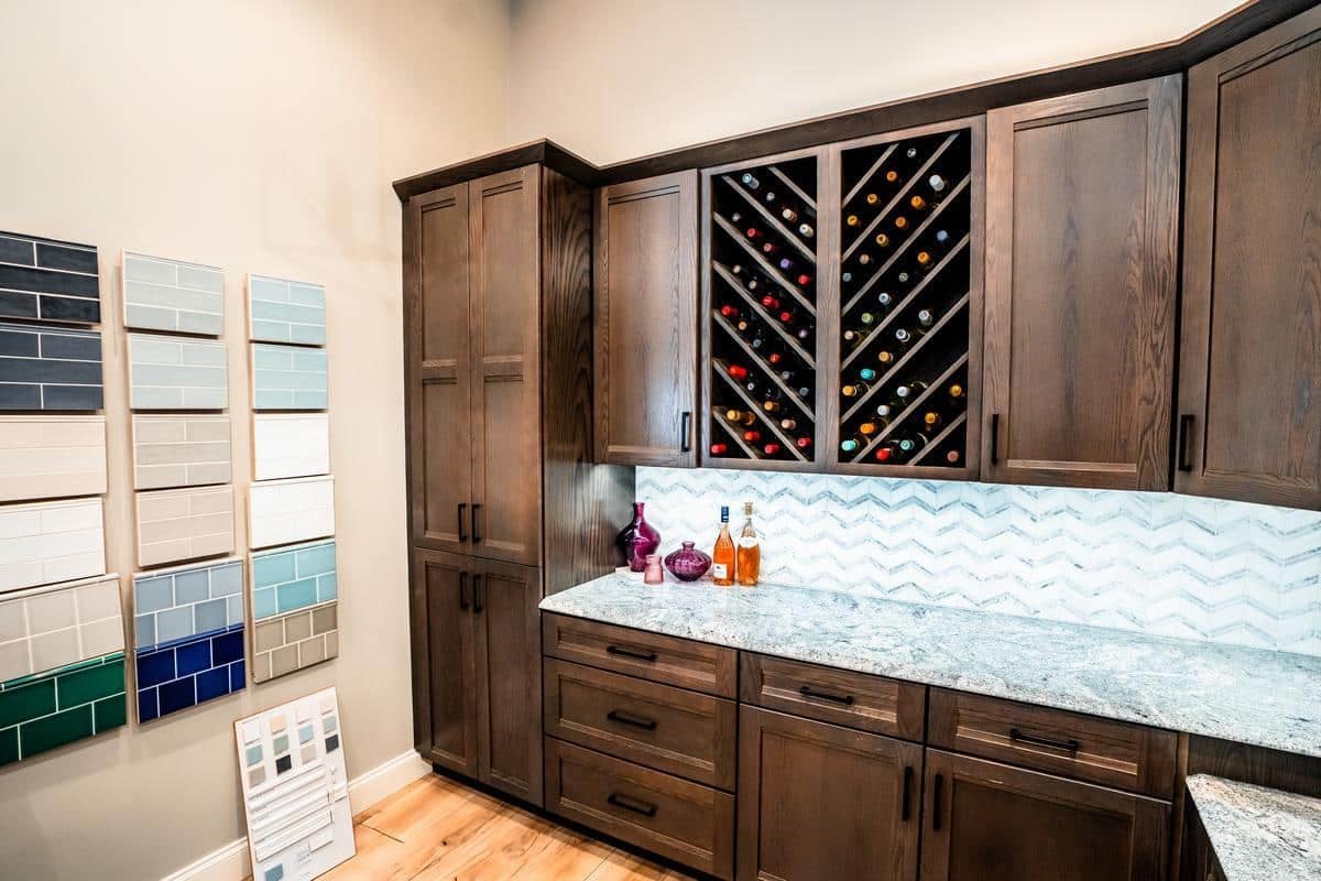 A modern kitchen at the Spencerport Showroom features dark wood cabinets, a granite countertop, a chevron-patterned tile backsplash, and a built-in wine rack. Colorful tile samples are displayed on the adjacent wall.