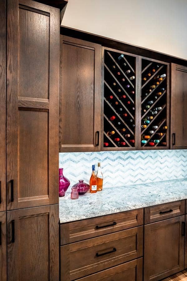 Modern kitchen showroom in Spencerport featuring dark wood cabinets, a granite countertop, chevron-patterned backsplash, wine bottles in a diagonal rack, and decorative glass bottles on the counter.
