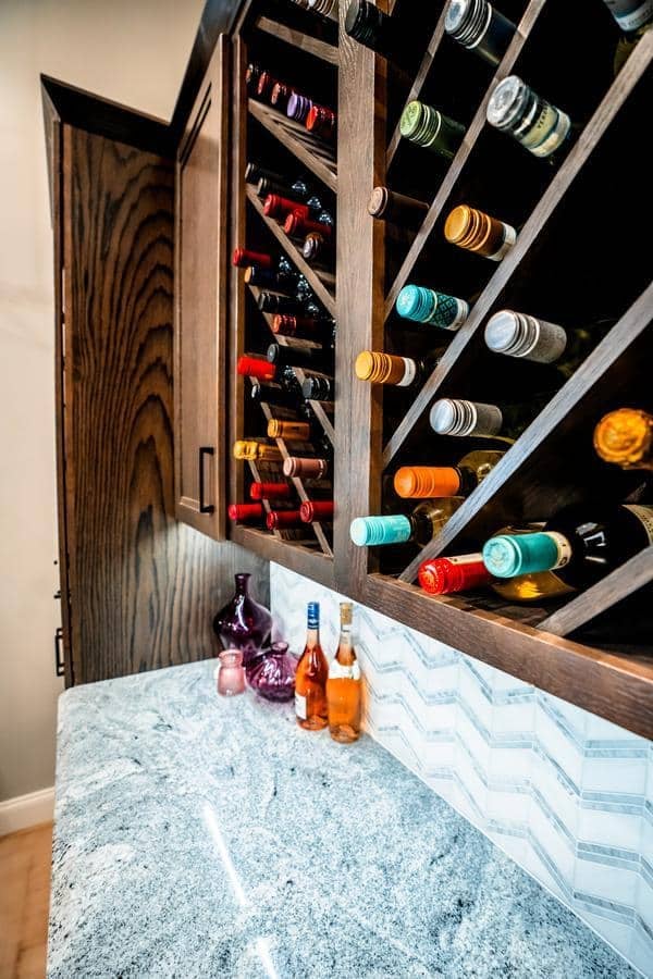 A modern kitchen countertop with a built-in wooden wine rack holding assorted wine bottles, plus several bottles and decorative glassware on the marble surface below, as seen in our Spencerport Showroom.