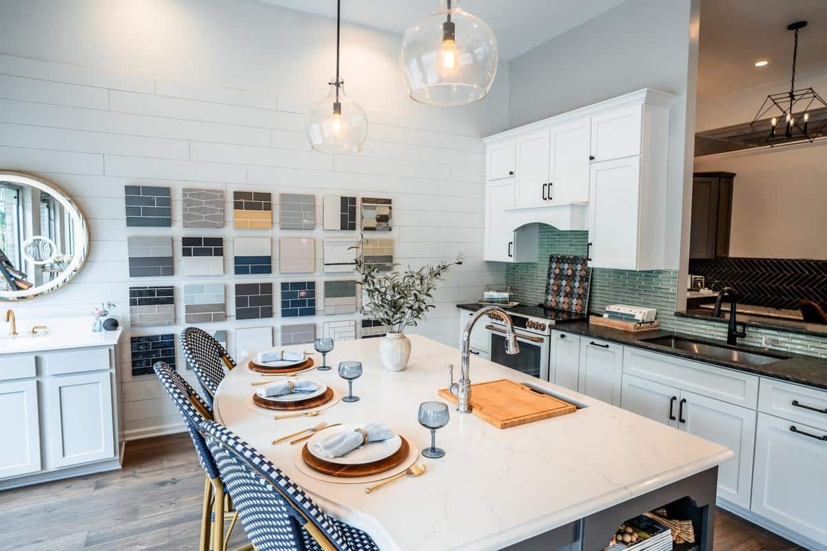 Modern kitchen with a white island, set for four with plates and gray glasses, rattan barstools, pendant lights, and a backsplash sample display on the wall—perfect inspiration from the Spencerport Showroom. Greenery in a vase serves as a centerpiece.
