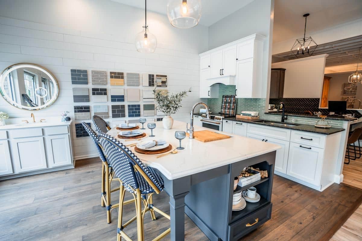 Modern kitchen with a large island featuring a sink and seating for four, white cabinetry, wooden floors, pendant lights, wall art, and decorative table settings—discover this look at our Spencerport Showroom.
