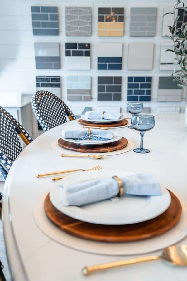 A modern dining table set with white plates, copper chargers, gold cutlery, and blue napkins, accompanied by blue glasses. Patterned chairs and a wall with tile samples from the Spencerport Showroom are in the background.
