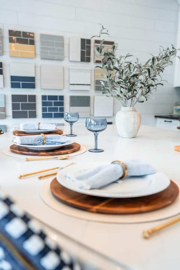 A modern dining table set with white plates, blue napkins, gold utensils, blue glasses, and a white vase with green branches is showcased in the Spencerport Showroom; color samples are displayed on the wall in the background.