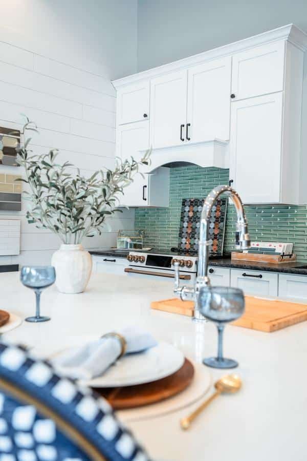 A modern kitchen with white cabinets, green glass tile backsplash, a stainless steel oven, and a white island countertop awaits in the Spencerport Showroom, featuring greenery, blue glassware, and gold utensils.