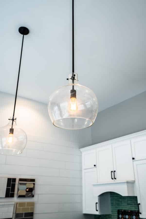 Two modern pendant lights with clear glass shades hang from the ceiling in a bright kitchen with white cabinets and light walls, showcasing stylish lighting designs available at the Spencerport Showroom. One bulb is turned on, illuminating the space.
