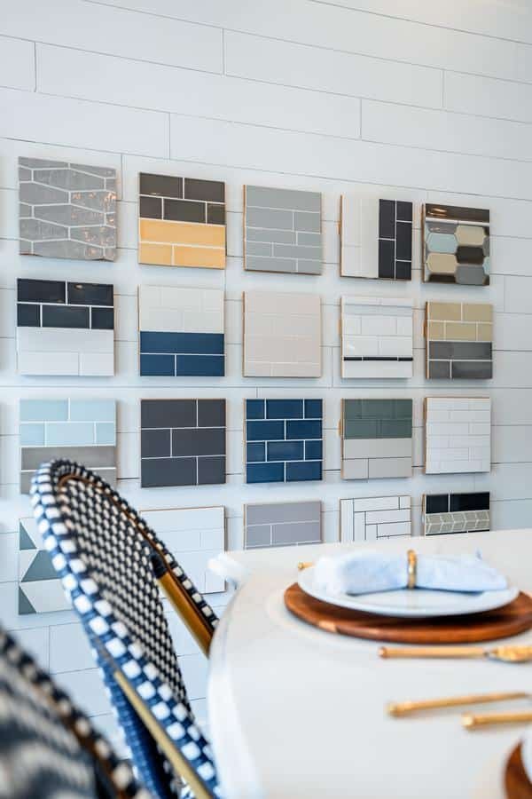 A display wall with various rectangular tile samples in different colors and patterns is featured in the Spencerport showroom, next to a table set with plates, napkins, and blue woven chairs.
