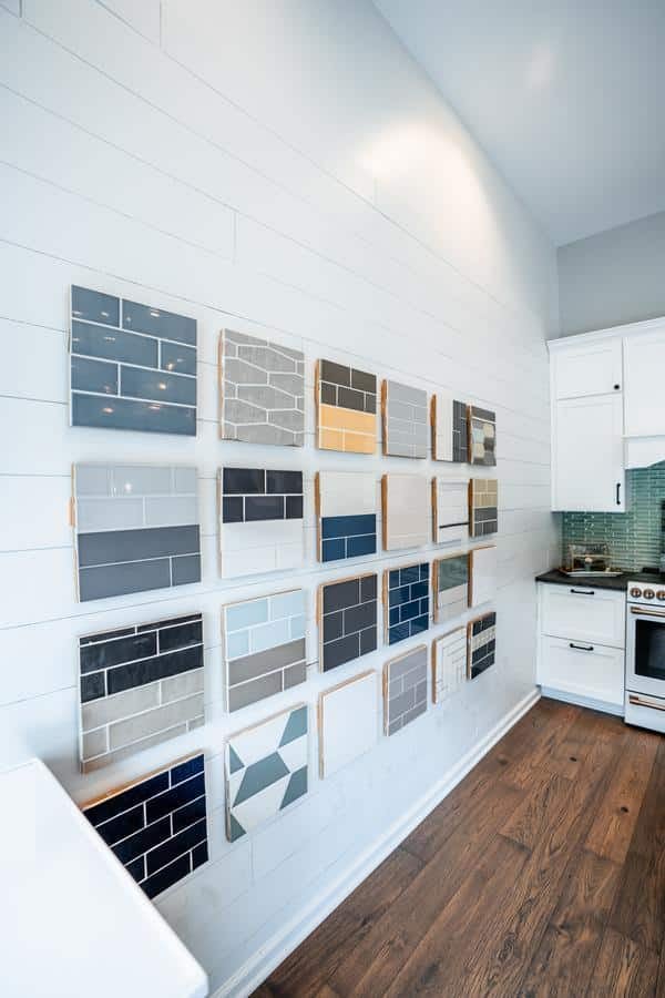Samples of various tile designs in different colors and patterns are displayed on a white shiplap wall in a modern Spencerport showroom with wooden floors and white cabinetry.