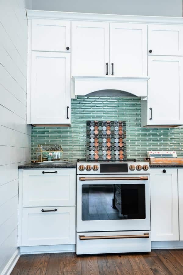 Modern kitchen with white cabinets, a black and copper stove, green glass tile backsplash, and wooden floor. Visit the Spencerport Showroom to see a decorative tray and cookbook displayed on the countertops.