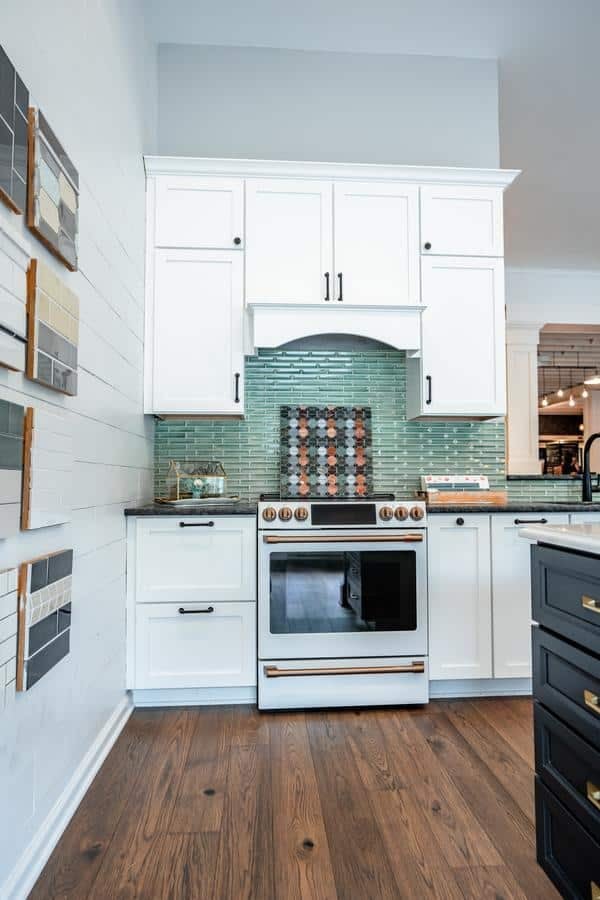 A modern kitchen showroom in Spencerport features white cabinets, a stainless steel oven, a green glass tile backsplash, wood flooring, and sample tiles displayed on the left wall.
