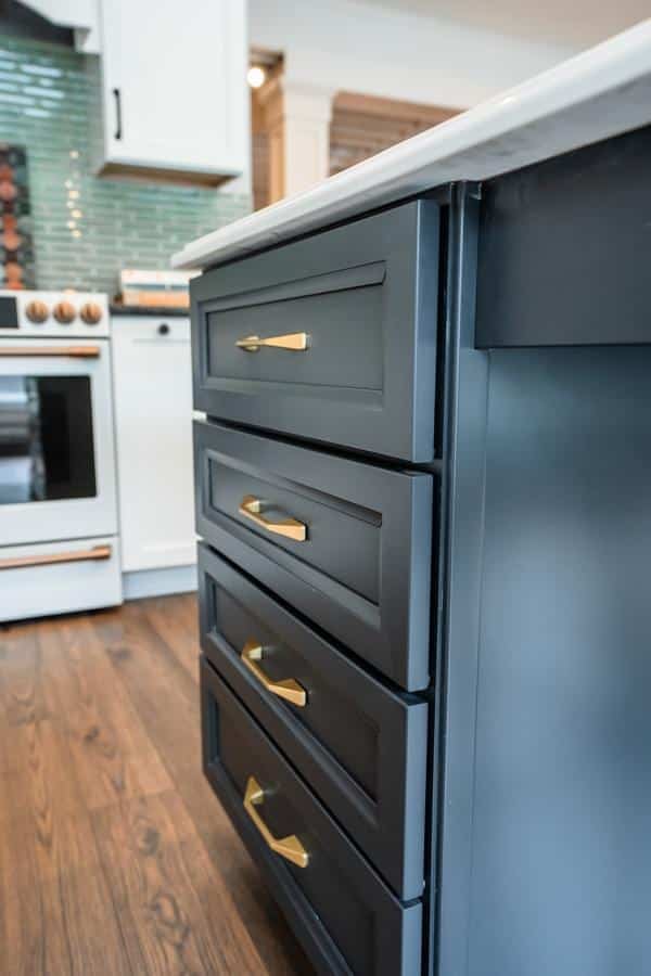 Close-up of a kitchen island with dark blue drawers and gold handles, next to a white stove and green tile backsplash on a wood floor in the Spencerport Showroom.