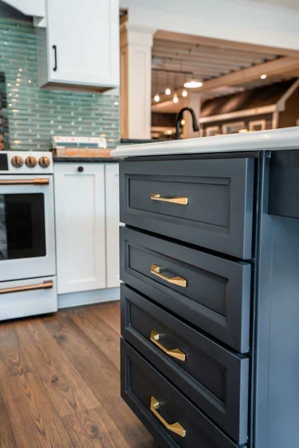A modern kitchen, available at the Spencerport Showroom, features black drawers with gold handles, white cabinets, a white oven, green subway tile backsplash, and wooden flooring.