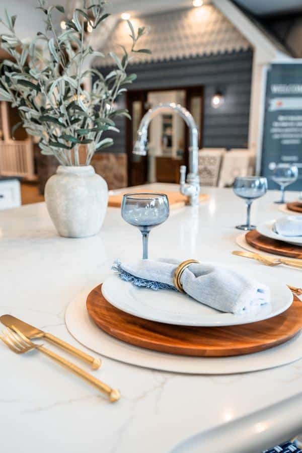 A close-up of a dining table set with a wooden charger, white plate, gold cutlery, and a blue-tinted wine glass in a Spencerport showroom. A napkin with a gold ring and greenery in a vase decorate the marble countertop.